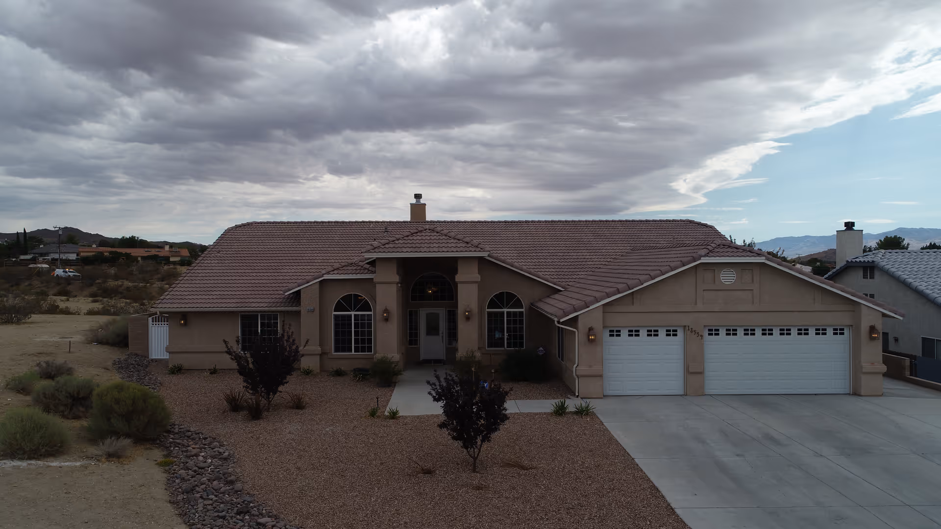 Front exterior view of a single-story house with a tiled roof, three-car garage, and a gravel yard with sparse desert landscaping under a cloudy sky.