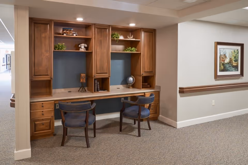 A hallway area in a senior living facility featuring a built-in wooden desk with two chairs. The desk has shelves with decorative items including a small globe, vintage camera, and plants. The walls are light-colored with a framed painting on the right side and a carpeted floor.