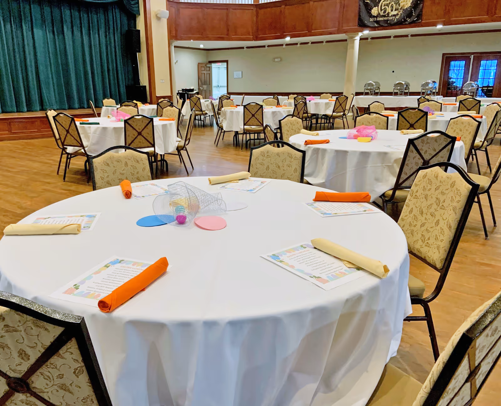A large event room with round tables covered in white tablecloths, each set with folded napkins in orange and beige, menus, and colorful centerpieces. The room has wooden floors, upholstered chairs, a stage with green curtains, and a balcony area above.