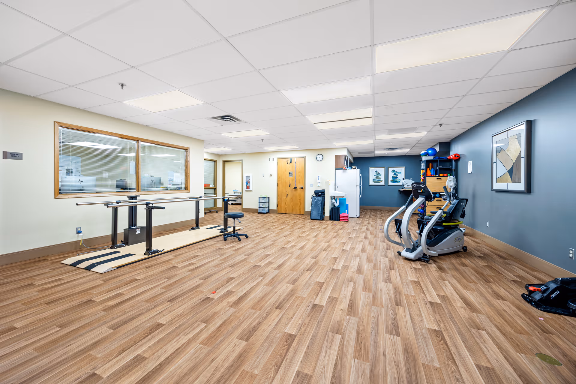 A spacious interior therapy/exercise room with parallel walking bars, exercise machines, and therapy equipment on wood-look flooring.