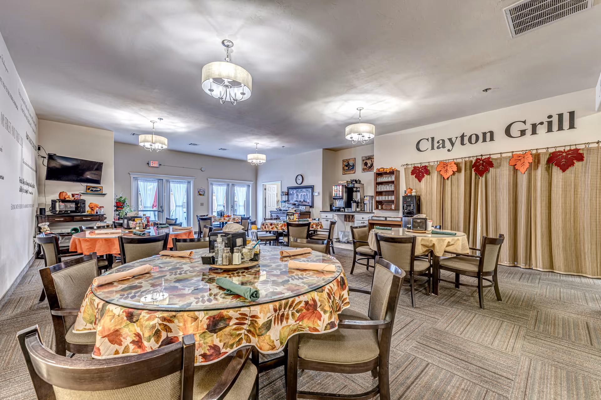 Dining area named Clayton Grill with multiple round tables covered with autumn-themed tablecloths, set with napkins and condiments. The room has several chairs around the tables, ceiling lights, a TV mounted on the wall, and a beverage station in the background. The decor includes fall leaf decorations hanging above a curtained area.