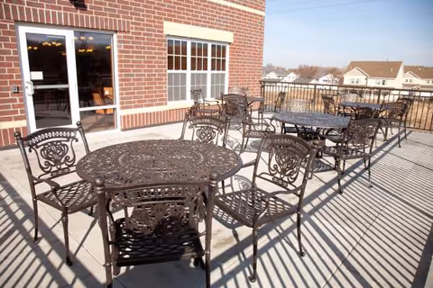 Outdoor patio area with multiple ornate metal tables and chairs arranged on a concrete surface next to a brick building with windows and a door. The area is fenced with a metal railing and residential houses are visible in the background under a clear sky.