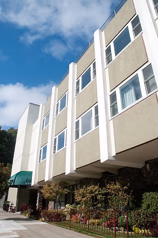 A multi-story beige senior living building with windows, greenery, and a green awning.
