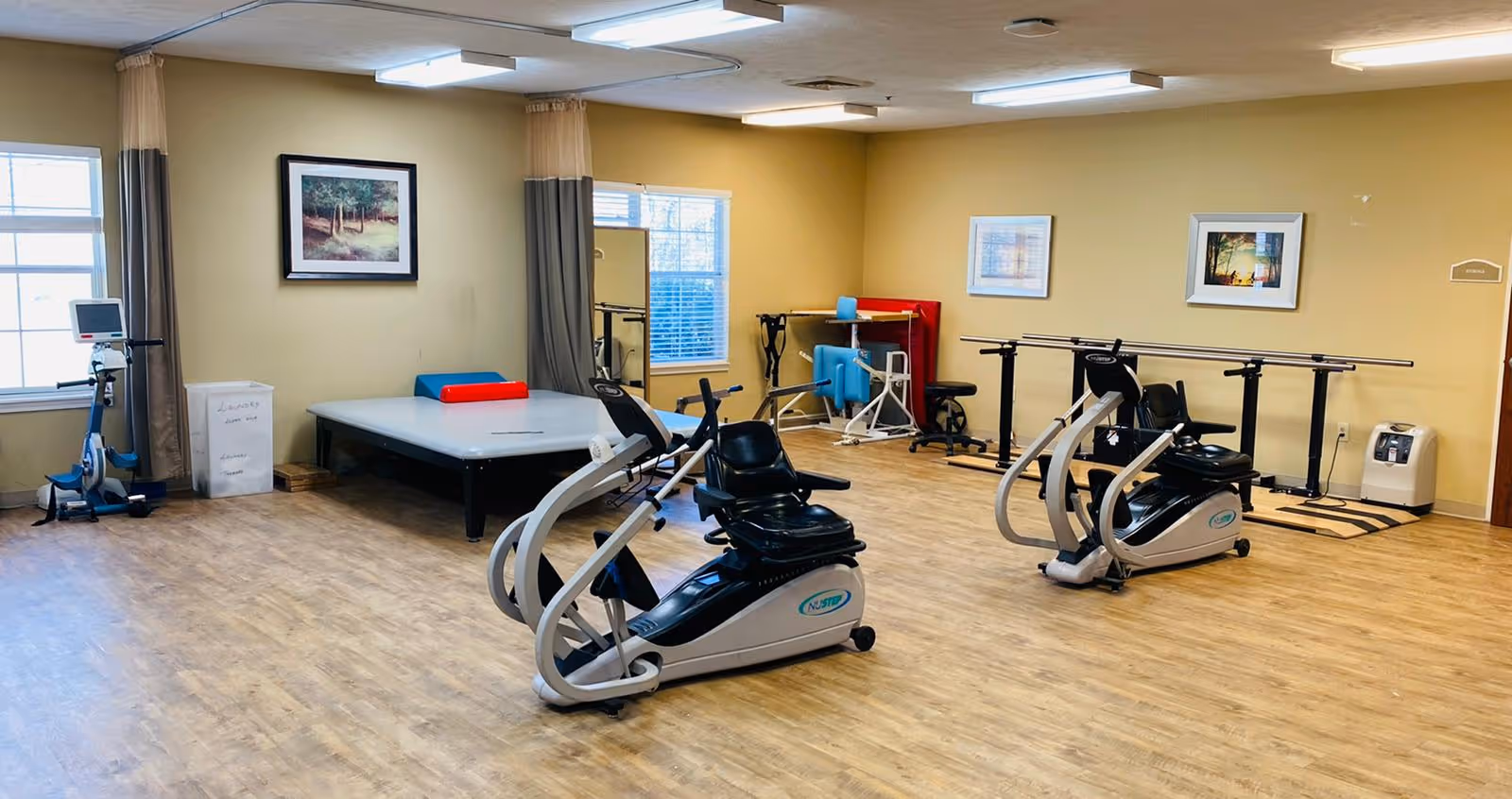 A rehabilitation room with exercise equipment including two recumbent exercise bikes, a stationary bike, parallel bars for walking exercises, and a padded therapy table. The room has wooden flooring, beige walls, two windows with curtains, and framed pictures on the walls.