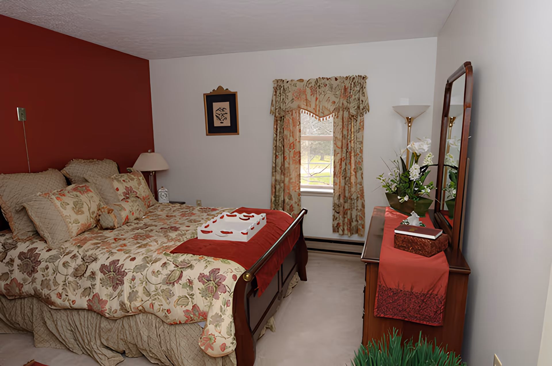 Cozy bedroom with a floral-patterned bed, wooden dresser and mirror, window with floral curtains, and decorative lamps and plants.