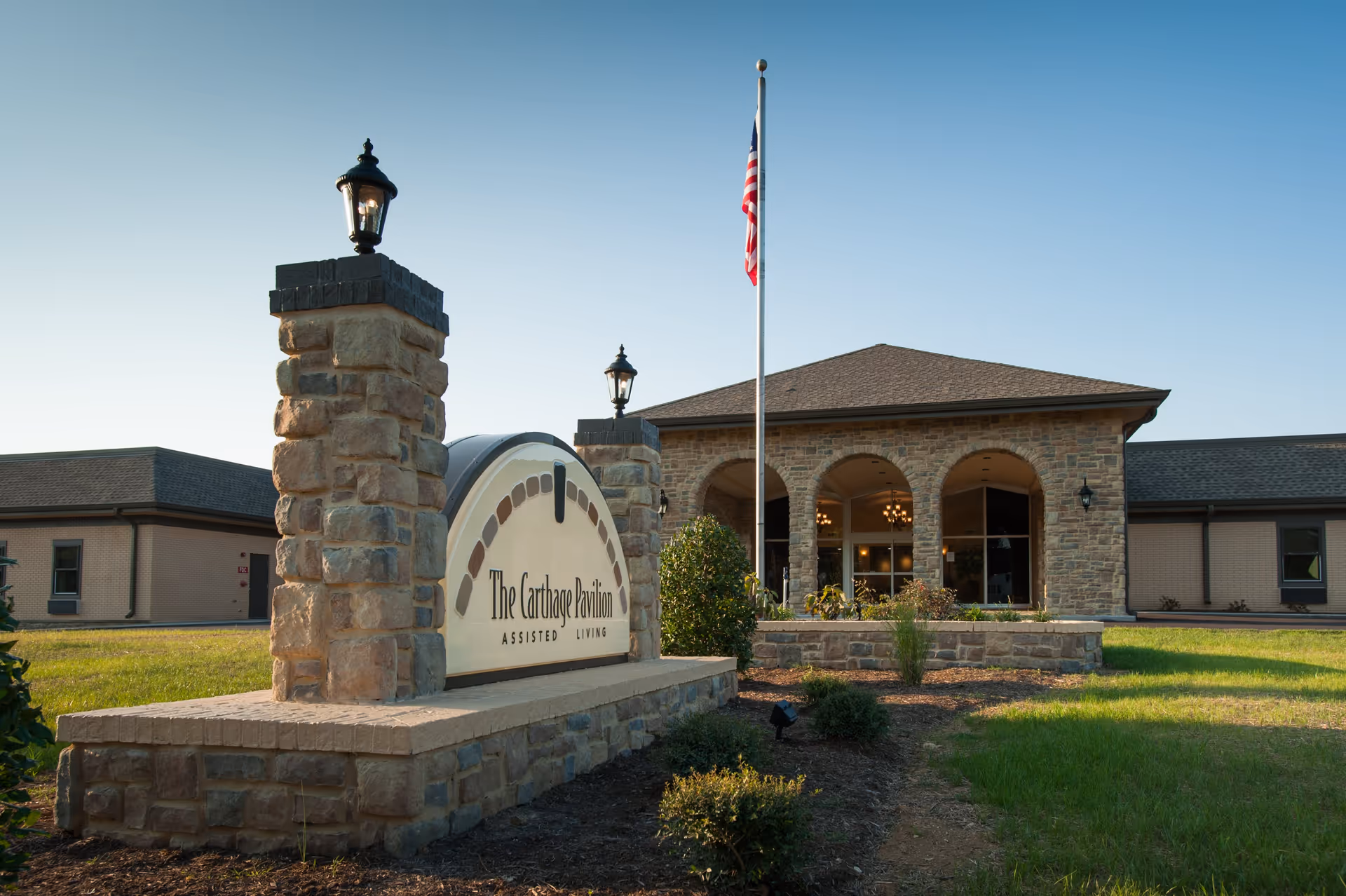 Exterior view of The Carthage Pavilion Assisted Living facility showing a stone sign with the facility name, a flagpole with an American flag, and a building with arched windows and stone facade under a clear sky.