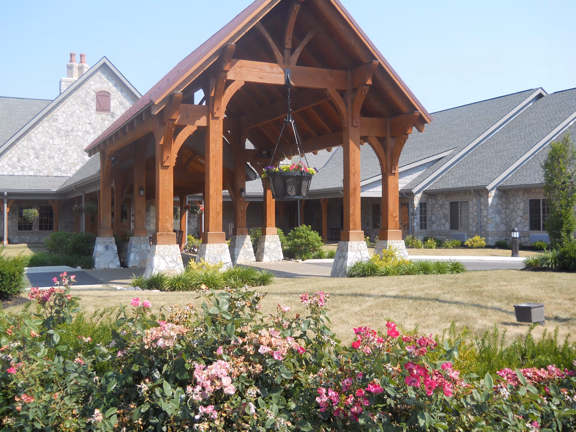 Stone building with a large wooden covered entrance supported by stone pillars, surrounded by a lawn and blooming pink flowers in the foreground under a clear blue sky.
