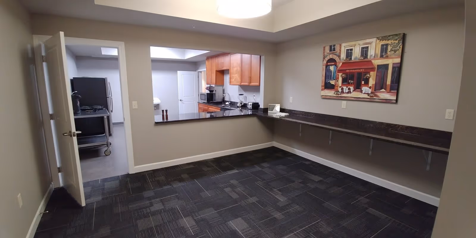 Empty room with dark patterned carpet and a long wall-mounted counter. The room has a large pass-through window opening to a kitchen area with wooden cabinets, a microwave, and a refrigerator visible. A painting of a restaurant scene hangs on the wall above the counter.