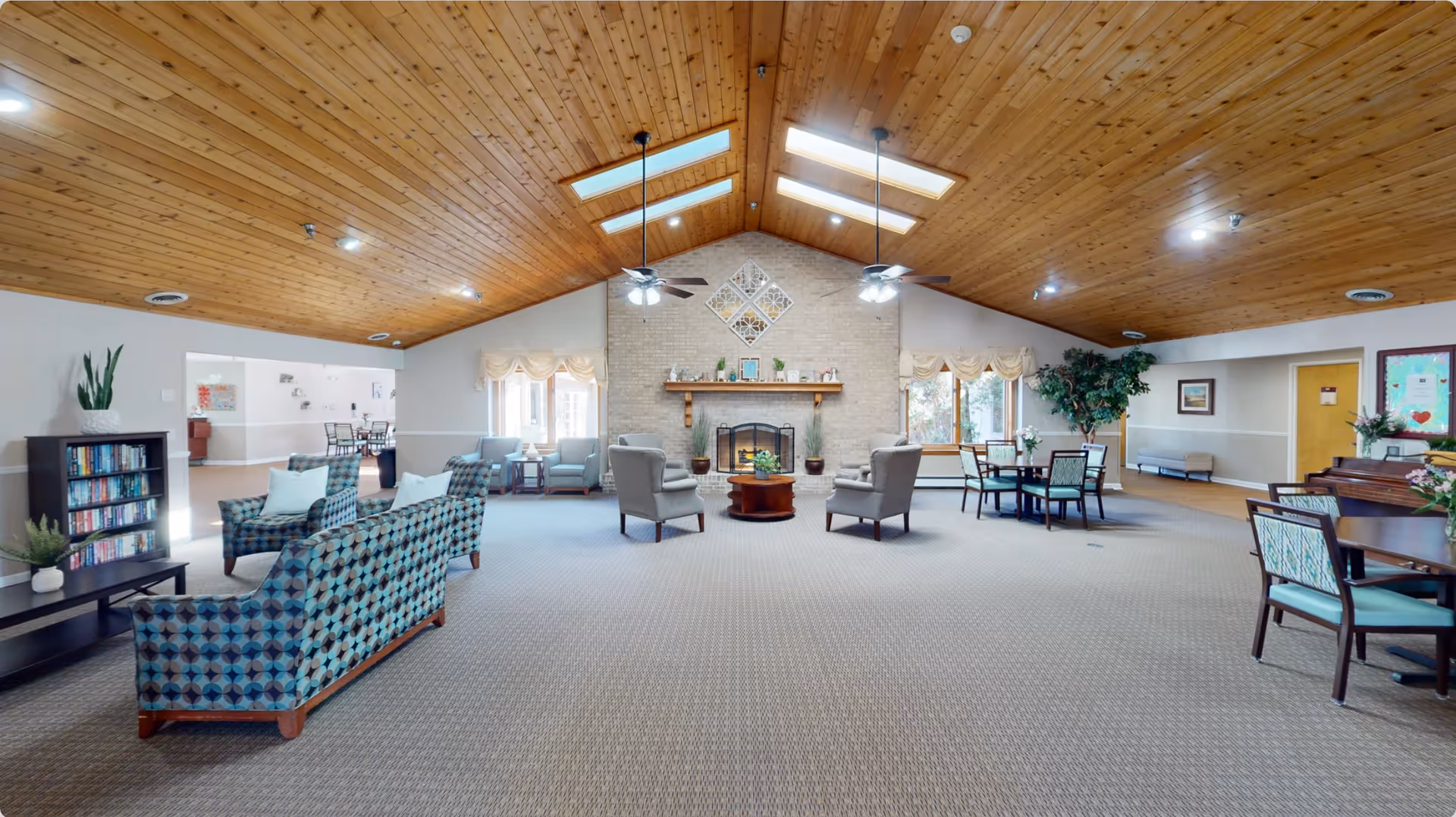 Spacious assisted-living common room with a vaulted wood ceiling, skylights, central fireplace, and seating and dining areas.