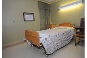 A simple nursing home bedroom with a single hospital-style bed covered with a patterned bedspread. The room has light green walls, a window with green curtains, a small wooden cabinet, and a chair next to the bed. There is a framed picture on the wall and a fluorescent light fixture above the bed.