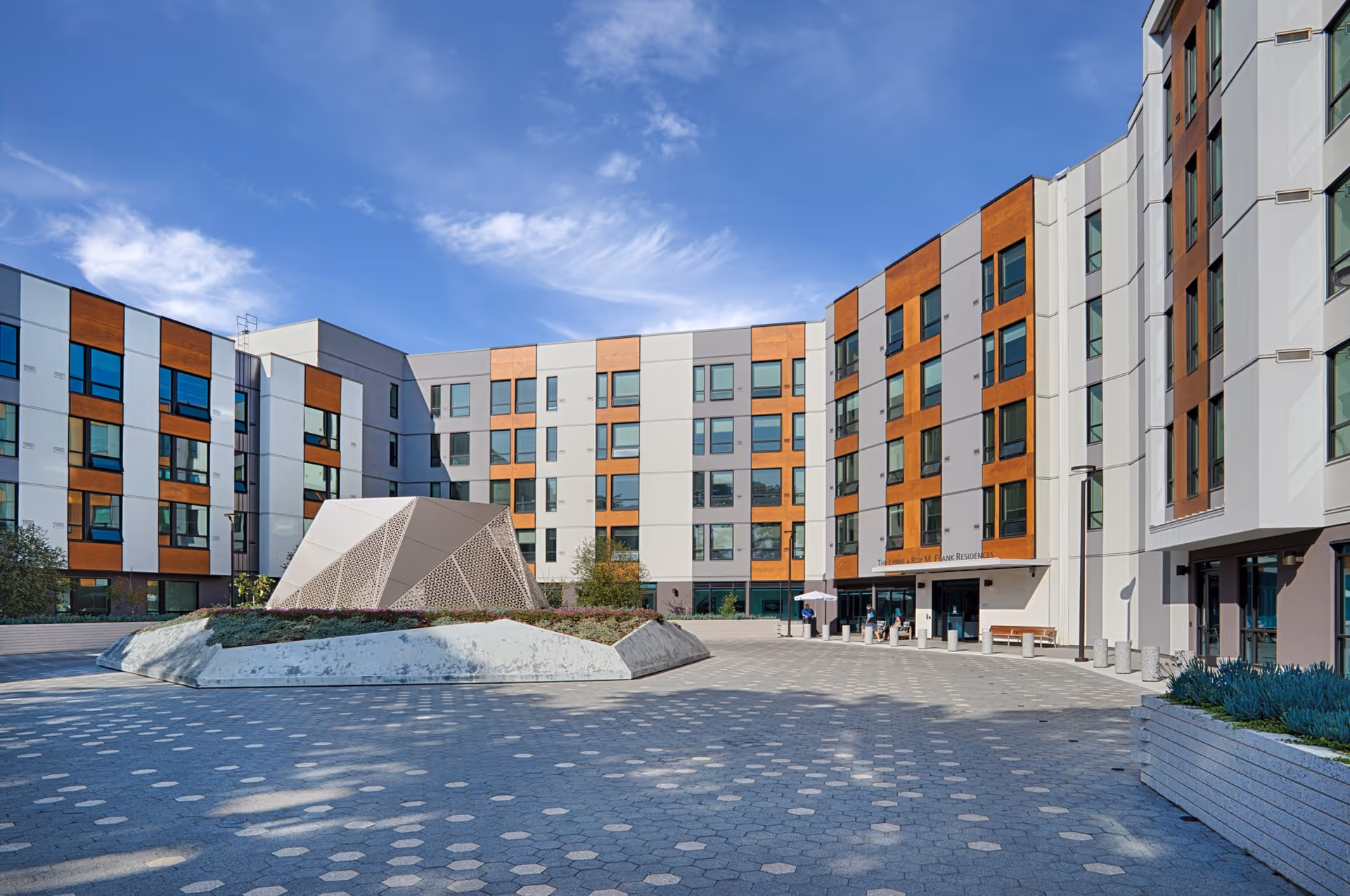 Exterior courtyard view of Frank Residences - Assisted Living & Memory Care, showing a modern multi-story building with white and wood-paneled sections, large windows, and a geometric sculpture in the center of the paved courtyard under a blue sky with some clouds.
