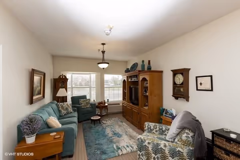 A cozy living room with a teal sofa, patterned armchair, and a wooden entertainment center holding a TV and decorative items. There is a grandfather clock on the wall, a small side table with a lamp, and a window with blinds letting in natural light. The room has a light-colored carpet and a blue and beige area rug.