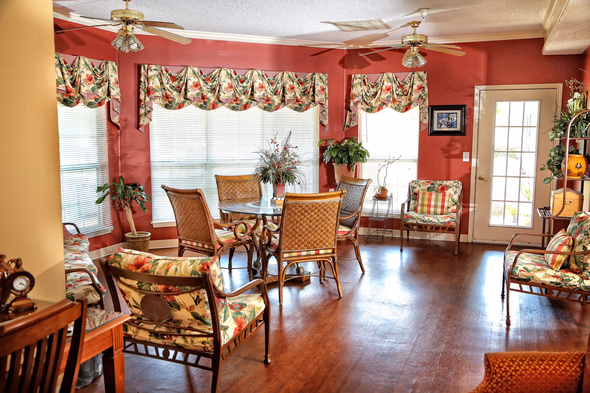 A bright communal living room with wicker chairs and floral cushions around a round glass table, red walls, hardwood floors, and large windows.