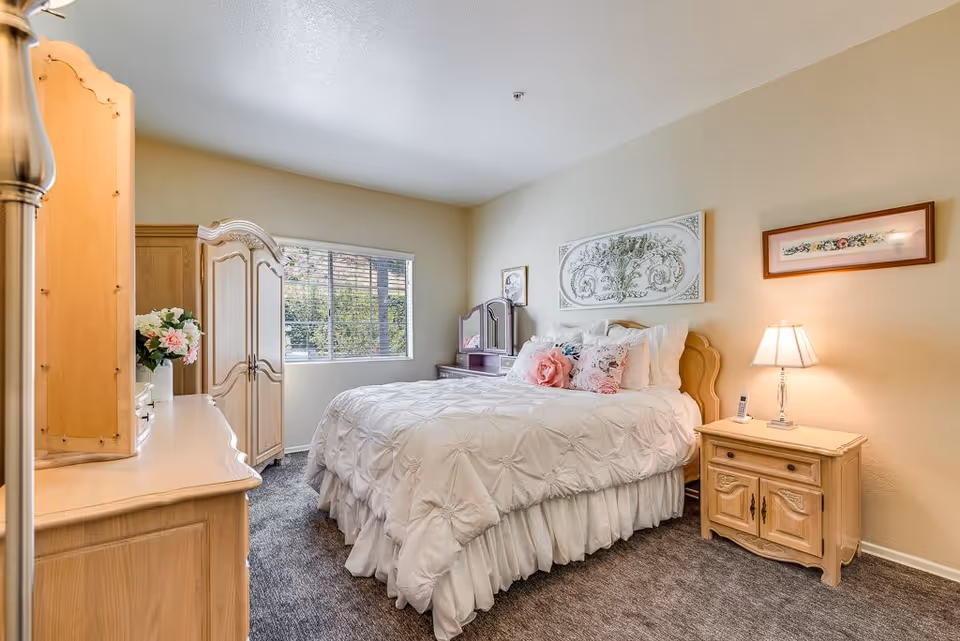 A cozy bedroom with a large bed covered in a white comforter and decorative pillows, a wooden nightstand with a lamp and a phone, a wooden dresser with a vase of flowers, a wooden armoire, and a window letting in natural light. The walls are decorated with framed artwork.