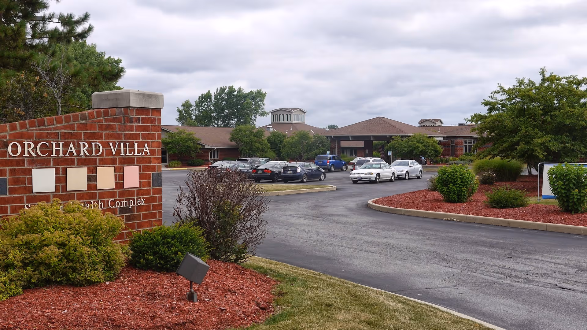 Exterior view of Orchard Villa senior living facility showing a brick sign with the facility name, a curved driveway leading to the building entrance, several parked cars, and landscaped areas with bushes and trees under a cloudy sky.