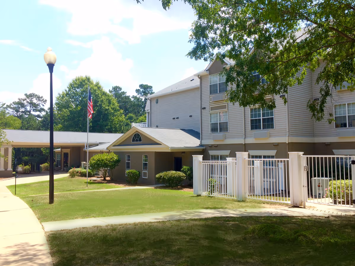Exterior view of a senior living facility building with beige siding and multiple windows, surrounded by green trees and bushes. There is a white fence in front, a sidewalk, a street lamp, and an American flag on a flagpole near the entrance.