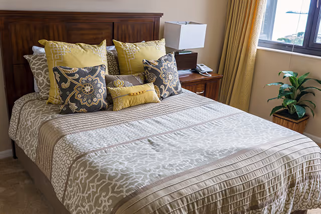 A neatly made bed with a wooden headboard, adorned with multiple decorative pillows in yellow, gray, and beige patterns. Next to the bed is a wooden nightstand with a modern white lamp and a telephone. A window with yellow curtains and a potted plant on the floor are visible in the background.