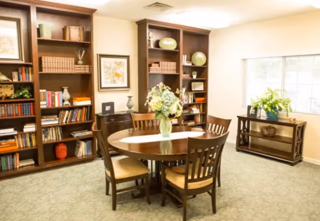 Bright communal room with a round wooden dining table and chairs, bookshelves, and decorative plants by a window.