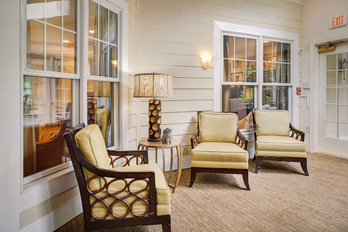 A cozy seating area inside a senior living facility with three cushioned armchairs arranged around a small round side table holding a decorative lamp and a small sculpture. The walls are light-colored with horizontal paneling, and there are large windows reflecting another room. A door with an exit sign is visible on the right side.