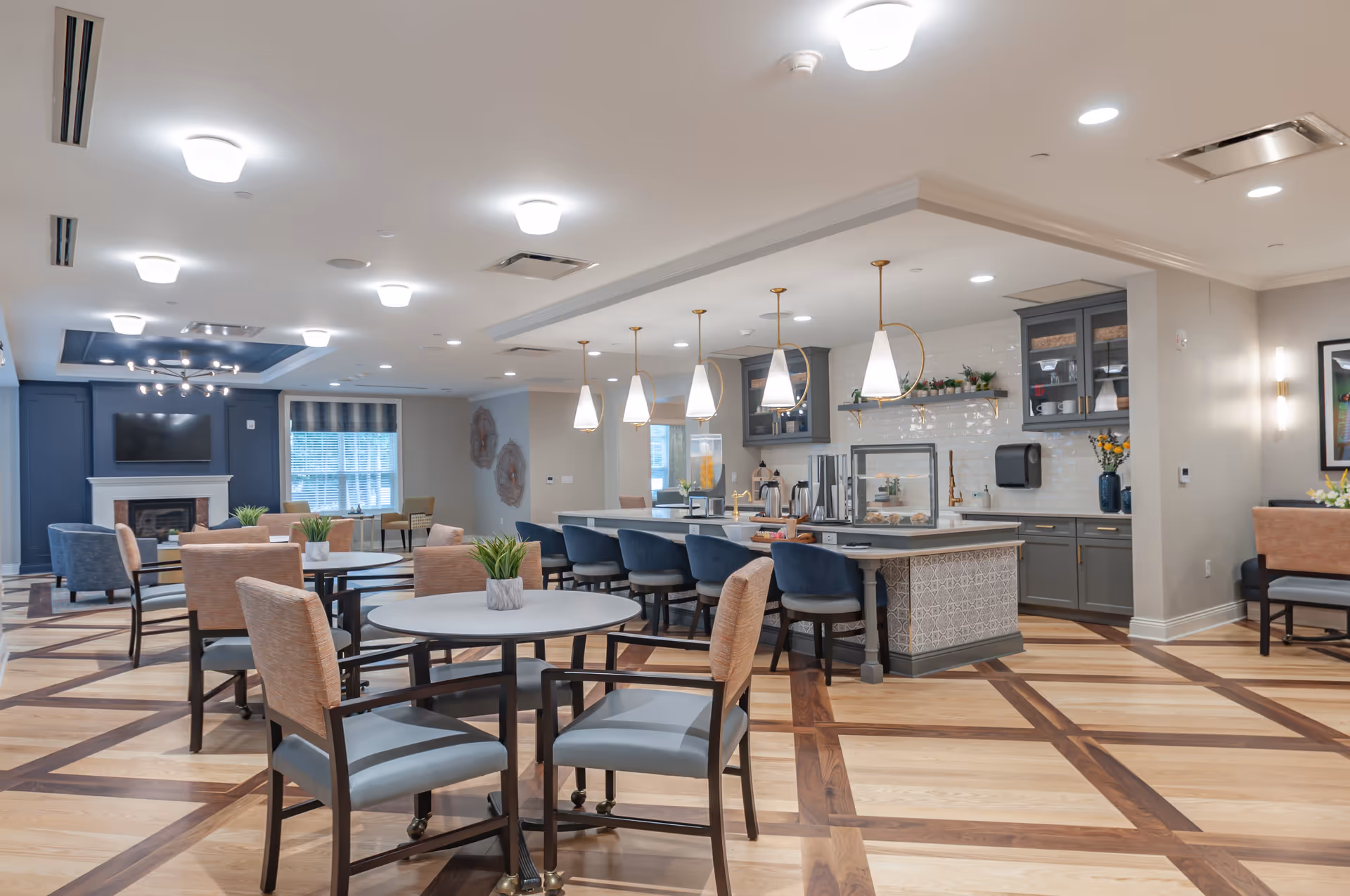 A spacious, well-lit common area in a senior living facility featuring multiple round tables with chairs, a kitchen island with bar stools, pendant lights hanging above the island, and a cozy seating area with a fireplace and television in the background.