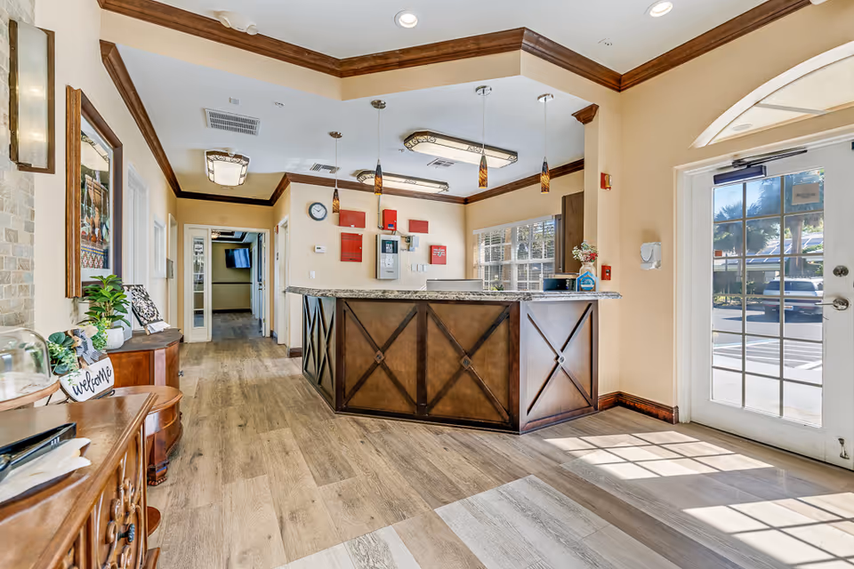 Reception area of Healthpark Senior Living featuring a wooden front desk with granite countertop, pendant lights hanging from the ceiling, a glass door allowing natural light, wooden flooring, and decorative plants and furniture along the walls.