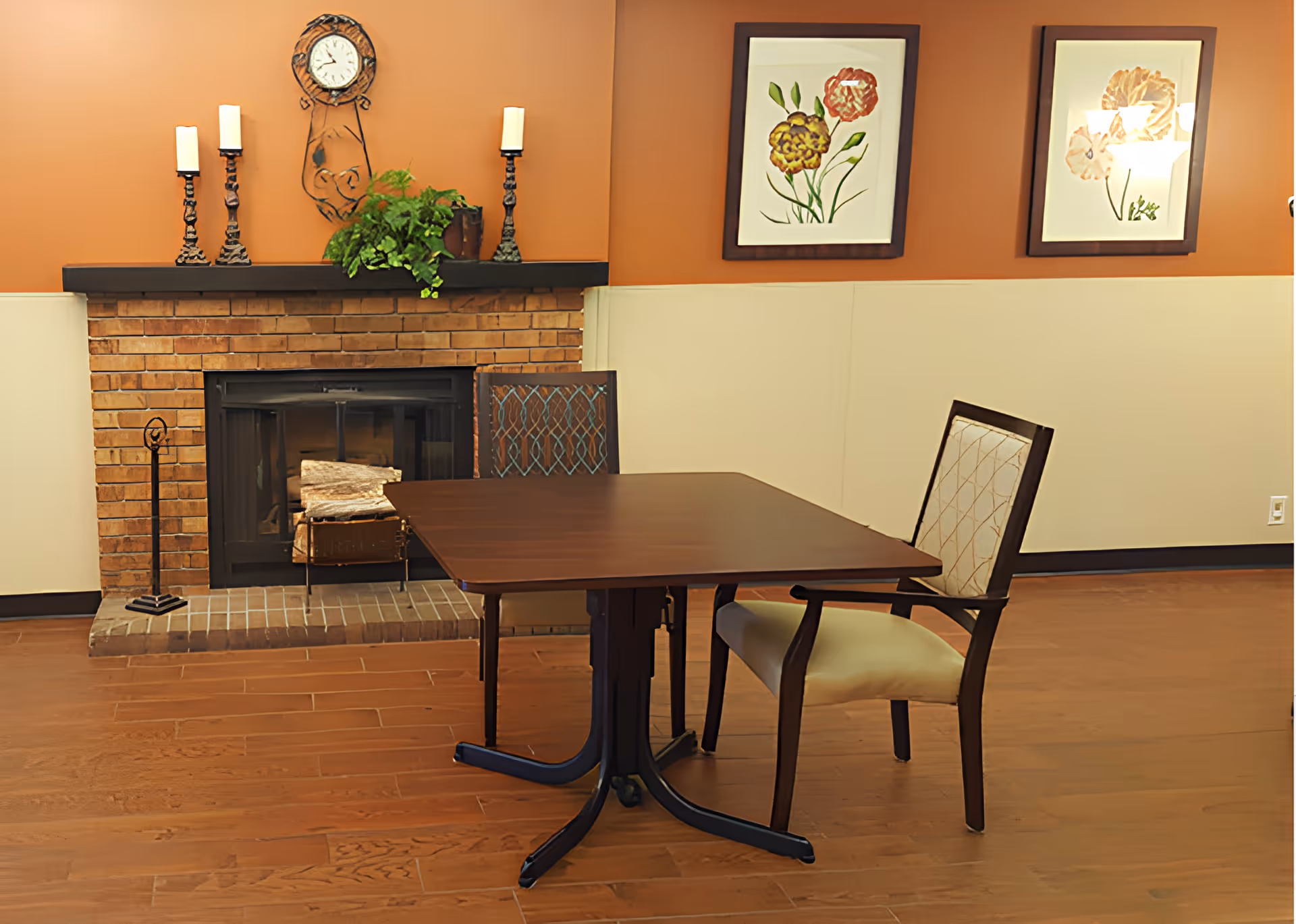 A cozy interior room with a brick fireplace, a wooden table, and two chairs. The fireplace mantle has three candles and a green plant. Two framed floral artworks hang on the orange wall above a beige wainscoting.