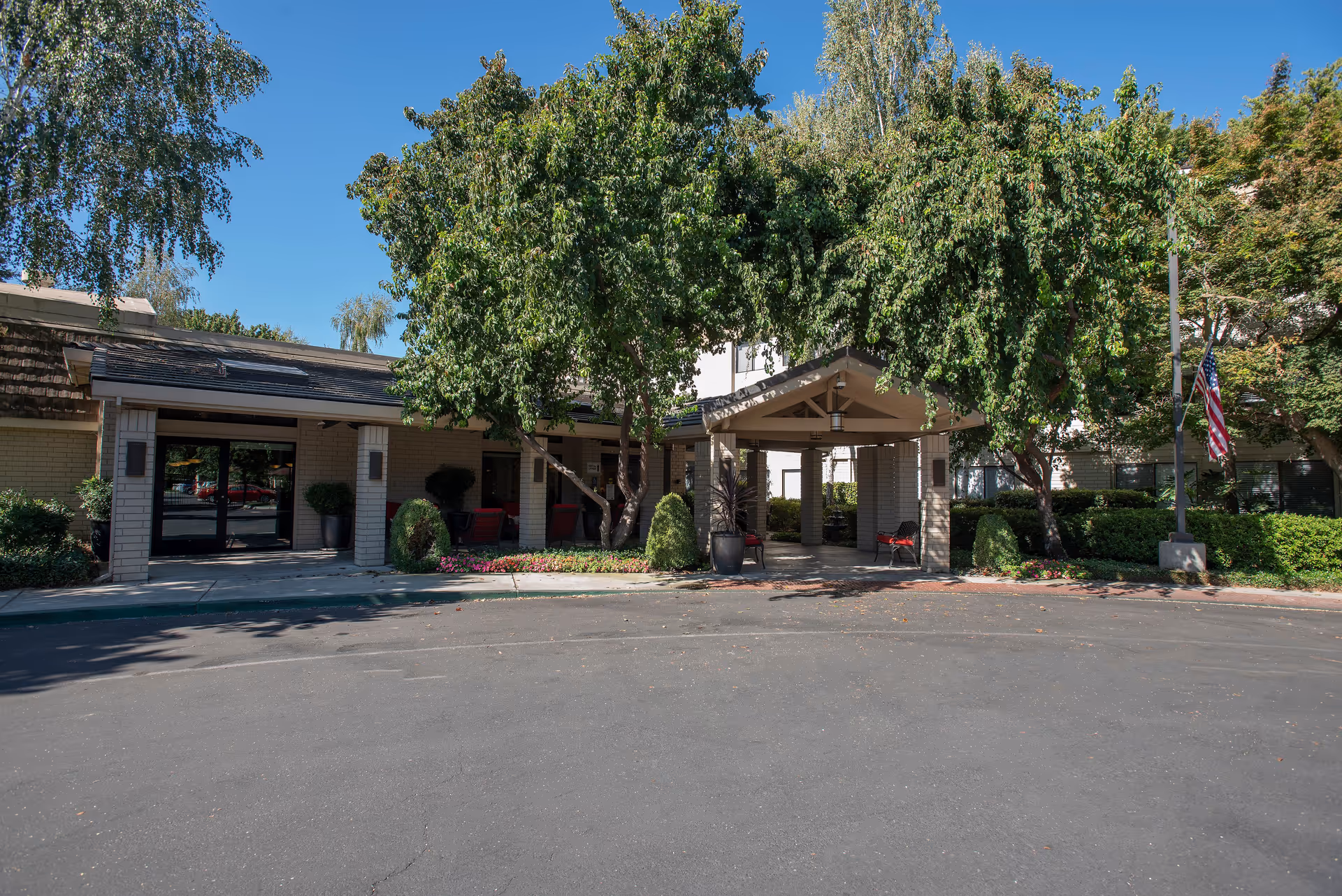 Exterior view of Campus Commons Senior Living facility entrance with a covered drop-off area, surrounded by lush green trees and shrubs under a clear blue sky. There are benches and potted plants near the entrance, and an American flag is visible on a pole to the right.