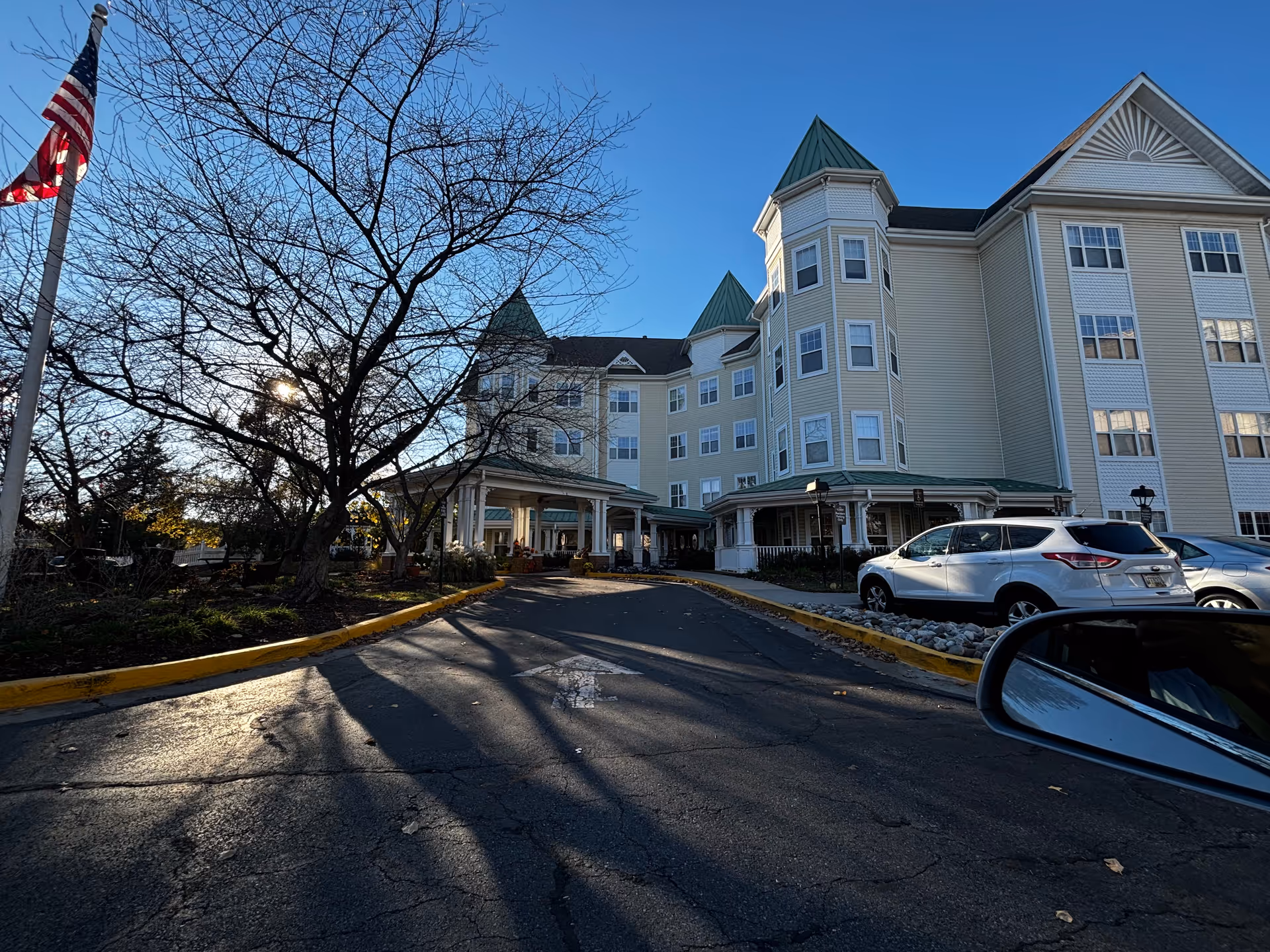 Front entrance and facade of a multi-story pale yellow senior living building with a leafless tree, parked cars, and an American flag.