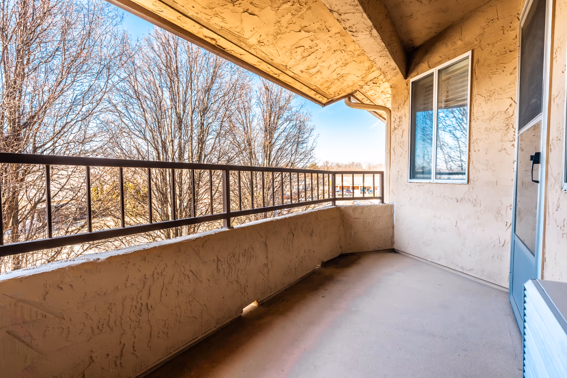 A covered outdoor balcony area with textured beige walls, a metal railing, a window, and a door. Leafless trees and a clear blue sky are visible in the background.