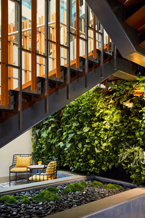 Indoor seating area with two chairs featuring yellow patterned cushions and a small round table, situated next to a lush green vertical garden wall under a modern staircase with wooden and metal railings. The floor has a decorative stone and moss arrangement.