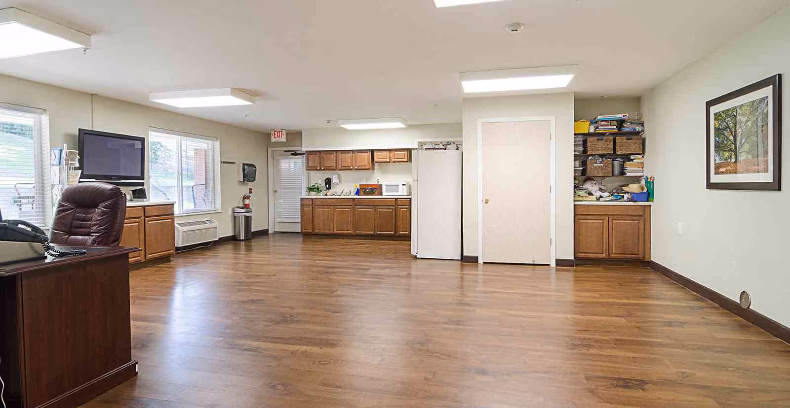 A spacious room with wooden flooring featuring a kitchenette area with wooden cabinets, a refrigerator, microwave, and a sink. There is a desk with a brown leather chair and a telephone on the left side, a wall-mounted TV, and a framed picture on the right wall. The room is well-lit with ceiling lights and has windows letting in natural light.