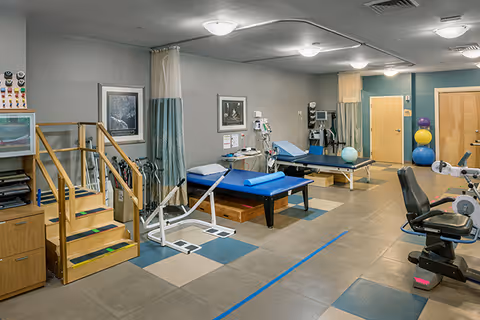 A physical therapy room with exercise equipment including parallel bars, therapy tables, exercise balls, and a stationary exercise bike. The room has tiled flooring with a mix of beige and blue tiles, gray walls, and ceiling lights. There are wooden doors and framed pictures on the walls.