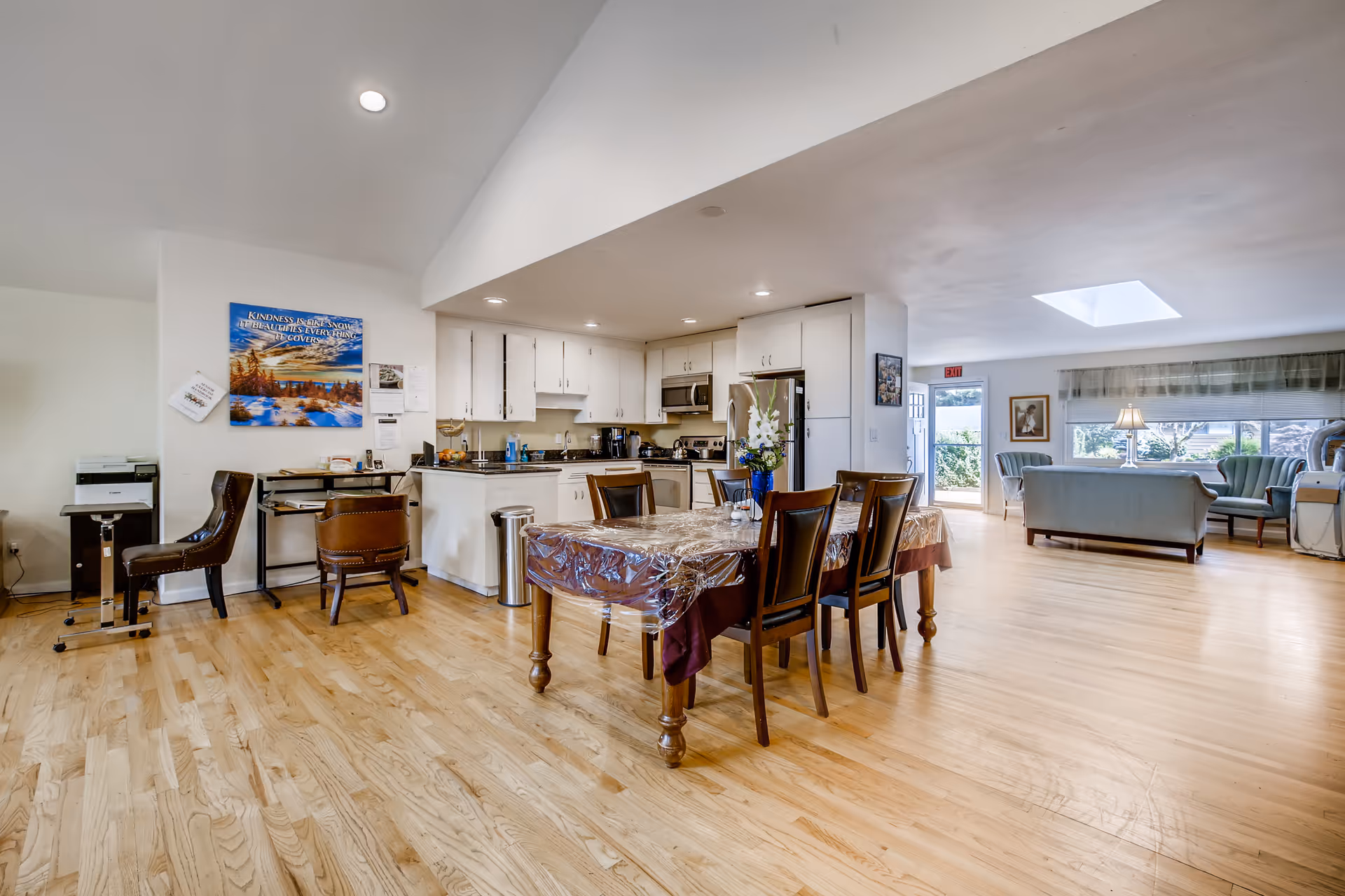A spacious, open-plan interior of a senior living facility featuring a kitchen with white cabinets and stainless steel appliances, a dining table covered with a plastic tablecloth and surrounded by six wooden chairs, and a living area with light blue upholstered sofas and armchairs near a large window. The room has light wood flooring, recessed ceiling lights, a skylight, and a wall with a motivational poster and some office equipment.