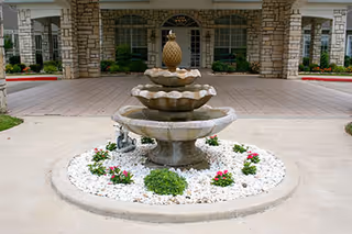 Stone Brook Assisted Living & Memory Care entrance featuring a tiered stone fountain with a pineapple finial, surrounded by white rocks and small flowering plants, with a covered driveway and stone pillars in the background.