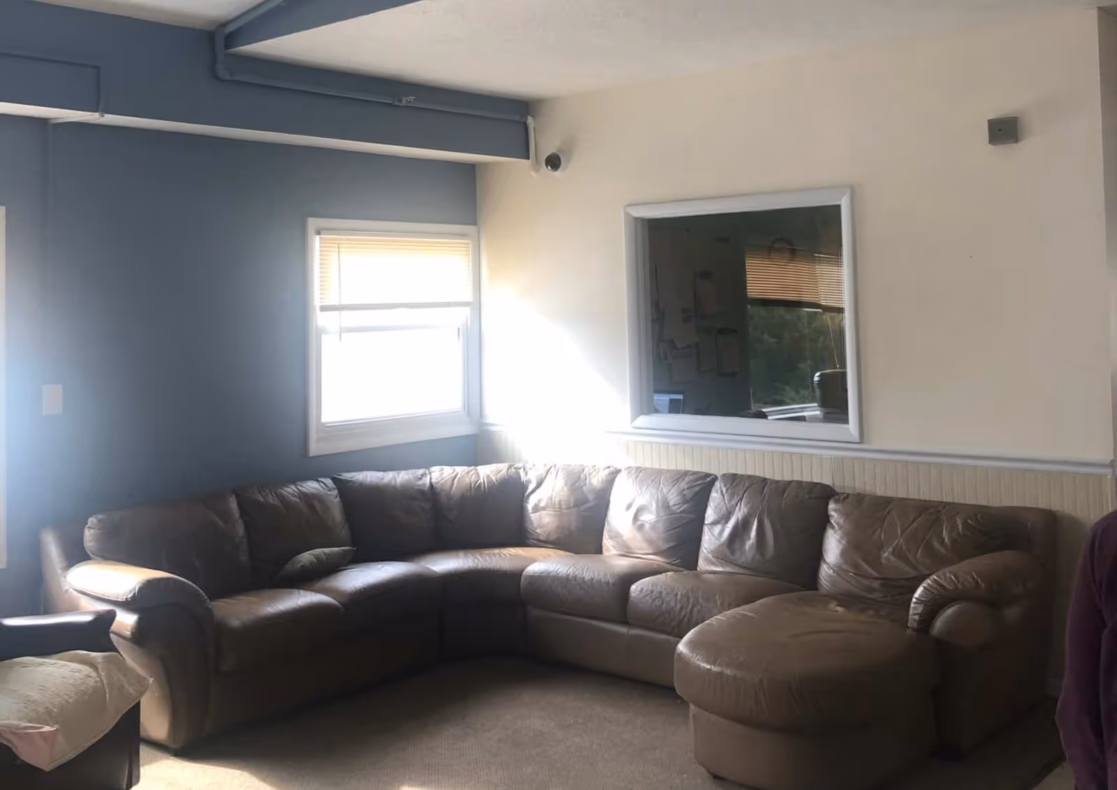 Sunlit common room featuring a large brown leather sectional sofa beneath windows and an interior pass-through window.