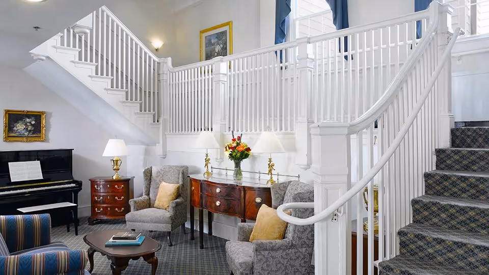 A cozy and elegant living area featuring a staircase with white railings and patterned carpet. The room includes two upholstered armchairs with yellow cushions, a small wooden coffee table with books, a wooden sideboard with three lamps and a vase of flowers, a piano with sheet music, and framed artwork on the walls.