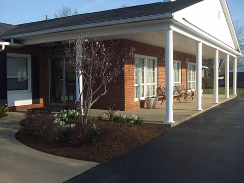 Exterior view of a single-story brick building with a covered porch supported by white columns. There are benches on the porch and a small landscaped area with a tree and plants near the entrance. The sky is clear and the driveway is paved.