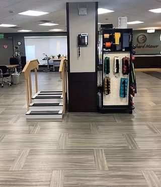 Interior view of a rehabilitation and nursing center showing a small set of stairs with handrails, a wall-mounted telephone, and a rack holding various physical therapy equipment such as resistance bands and weights. In the background, there is a reception desk with the sign 'Orchard Manor'.