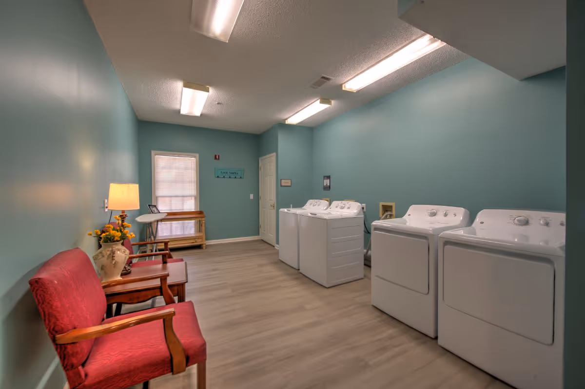Well-lit communal laundry room with multiple washers and dryers, teal walls, red chairs, and a folding table.