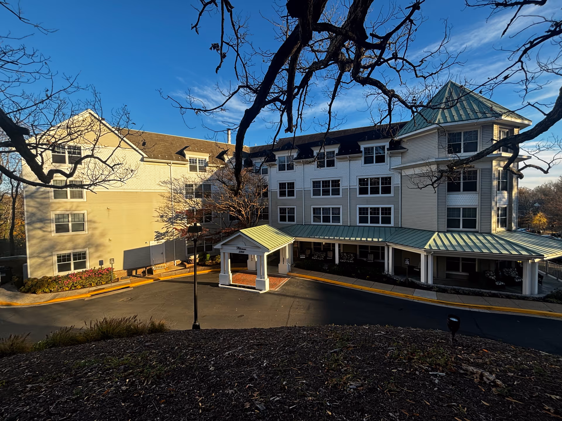 Exterior view of a multi-story senior living facility building with a covered entrance and green metal roofing. The building is surrounded by leafless trees and landscaping, under a clear blue sky.