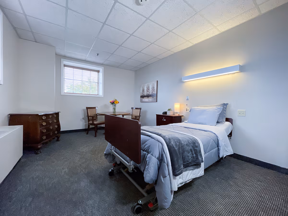 A simple, clean room with a single hospital-style bed made with gray and white bedding. There is a small wooden nightstand with a lamp and a small plant next to the bed. On the opposite side of the room, there is a wooden dresser and a small round table with two wooden chairs. A window with blinds lets in natural light, and a framed picture hangs on the wall above the nightstand. The room has a carpeted floor and a drop ceiling with fluorescent lighting.