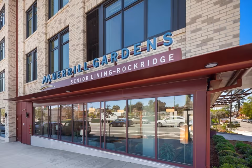 Exterior view of Merrill Gardens at Rockridge senior living facility showing the building's brick facade, large windows, and a maroon awning with the facility's name and 'Senior Living - Rockridge' signage above the entrance.