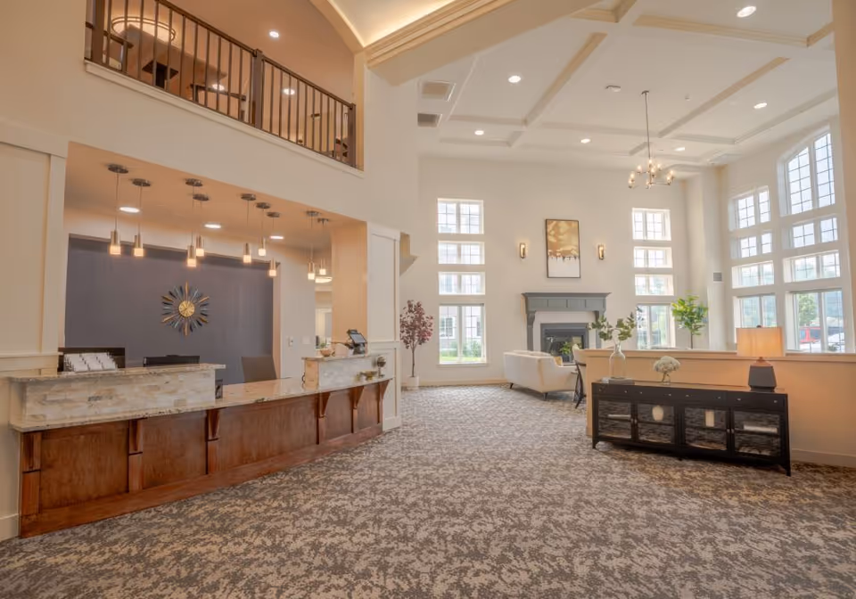 Spacious and well-lit senior living facility lobby with a reception desk on the left, a seating area with armchairs near a fireplace, large windows letting in natural light, and decorative plants and artwork.