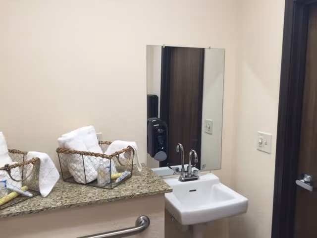 A small bathroom sink with a mirror above it, a soap dispenser mounted on the wall, a granite countertop with two wire baskets holding rolled white towels and toiletries, a handrail below the countertop, and a closed wooden door to the right.