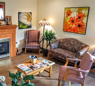 A cozy living room area with a floral patterned sofa, two armchairs, a wooden coffee table with magazines, a potted plant, a floor lamp, and colorful floral paintings on the walls. There is also a wooden fireplace on the left side.