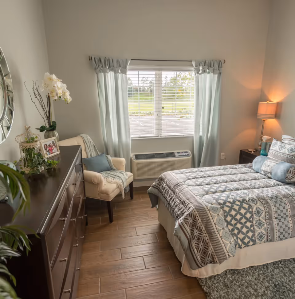 Neatly staged bedroom with a patterned bed, dresser, armchair, window with light blue curtains and a lamp on a wooden floor.