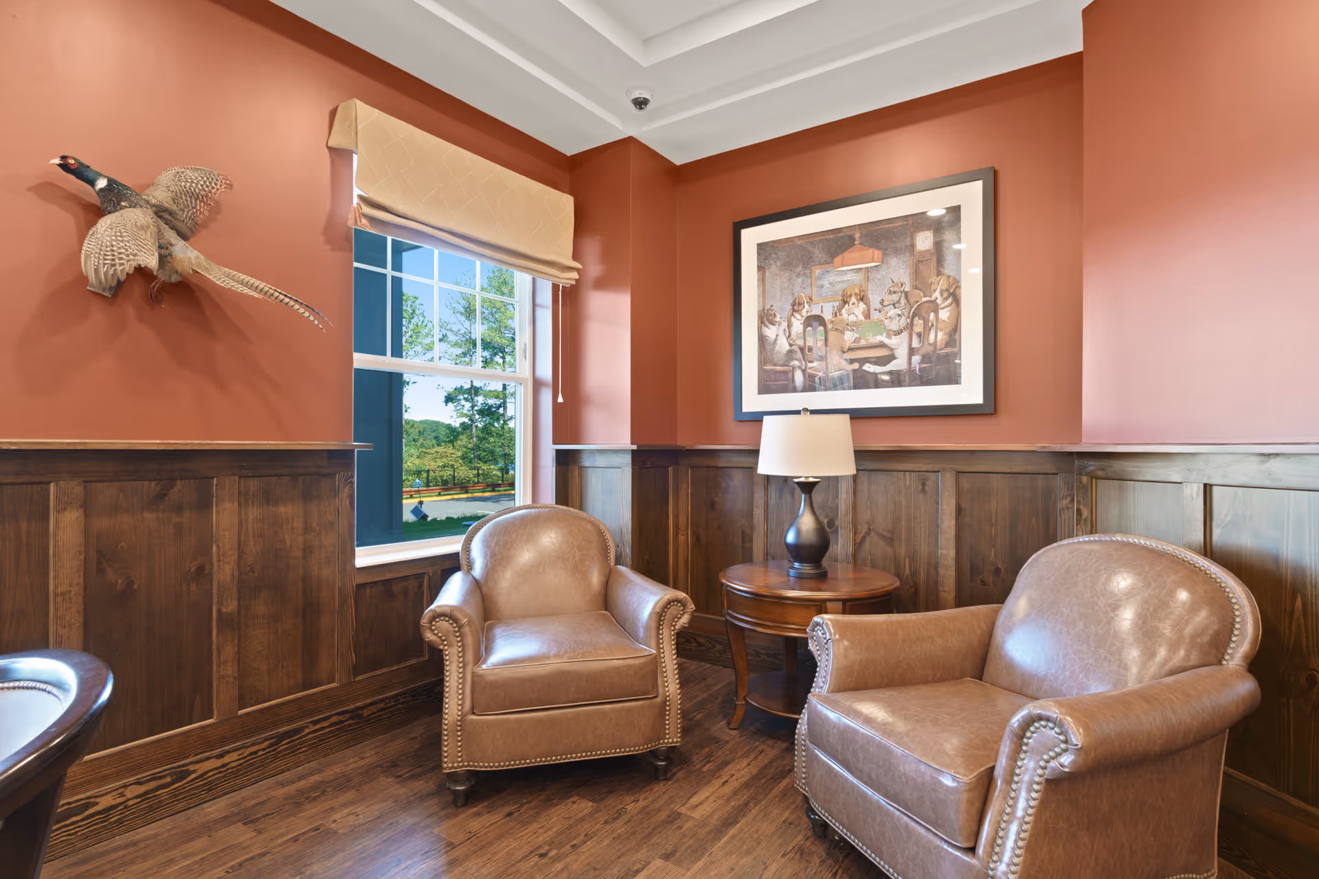 A cozy corner of a room with two brown leather armchairs, a round wooden side table with a lamp, a window with a beige roman shade, a mounted pheasant on the wall, and a framed painting of dogs playing poker on the wall with reddish-brown paint and wooden wainscoting.