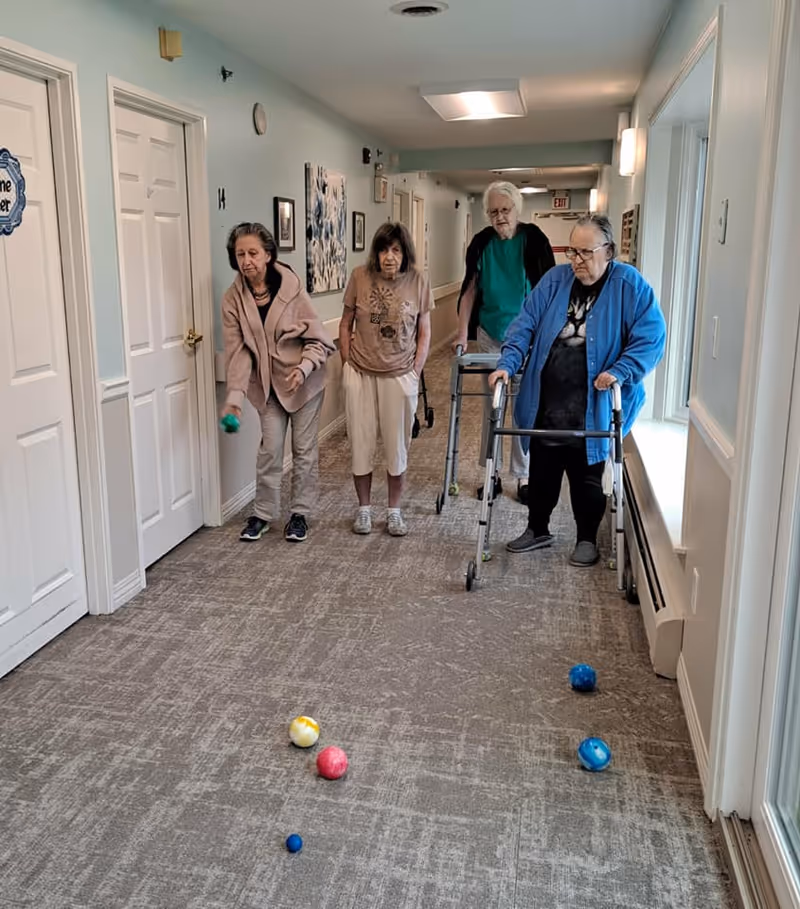 Four elderly individuals in a hallway of a senior living facility playing a game with colorful balls on the carpeted floor. One person is using a walker, and another is in the process of throwing a ball. The hallway has light blue walls, white doors, and windows on the right side letting in natural light.