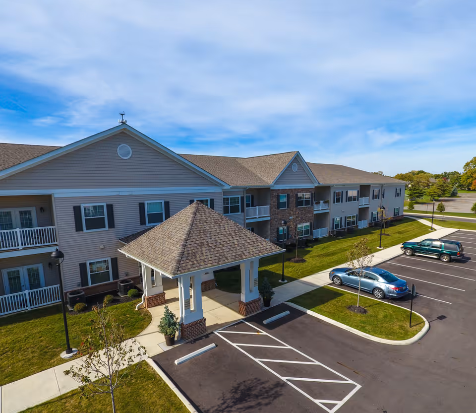 Front exterior of a two-story senior living building with a covered entrance, parking lot, and parked cars under a blue sky.