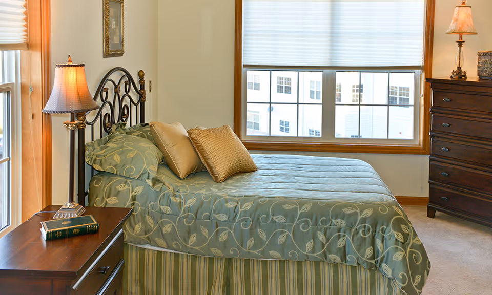 Bright bedroom with a patterned bed, decorative pillows, bedside tables and lamps next to a large window.